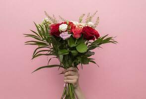Human Hand Holding Beautiful Fresh Bouquet Of Flowers For Bride Or Occasion On Pink Background. Colorful Mixed Roses, Carnation Shabot, Green Leaves, Gerber. Horizontal Plane. High quality photo