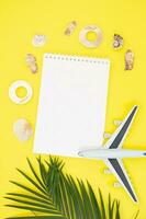 Plane with palm branches with frame of shells around notepad on summer yellow background. Vacation, travel concept, flat lay top view, minimal exotic concept. Verical. Copy space photo