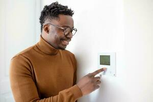 hombre afroamericano sonriente usando un moderno sistema doméstico inteligente, controlador en la pared, joven positivo cambiando la temperatura en el termostato o activando la alarma de seguridad en el apartamento foto