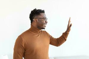 Smiling african American millennial man in glasses look at camera waving saying hello talking on video call, happy black male vlogger in spectacles greeting with subscribers shooting video blog photo