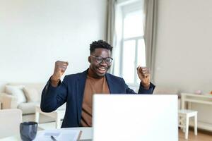 Excited African American man wearing headphones reading good news in email, getting new job, promotion, using laptop, looking at screen and screaming with joy, showing yes gesture, celebrating photo