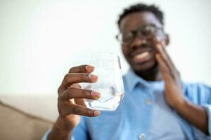 joven hombre con sensible dientes y mano participación vaso de frío agua con hielo. cuidado de la salud concepto. hombre Bebiendo frío beber, vaso lleno de hielo cubitos y siente dolor de muelas, dolor foto