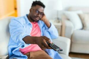 hermoso africano americano joven hombre sentado en el sofá a hogar, acecho televisión, relajante y mirando confuso foto