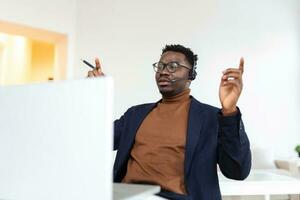 Smiling African American employee in headphones using laptop, looking at screen, making video call or watching webinar, writing notes, distance learning language concept, call center operator working photo