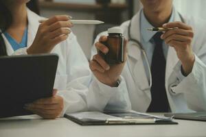 Confident doctor man holding a pill bottle and writing while talking with senior patient and reviewing his medication at office room. photo
