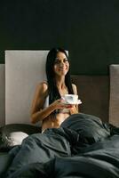 joven hermosa morena en pijama Bebiendo café sentado en cama en el Mañana después dormido foto