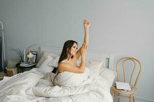 Pretty young woman is sitting on the bed wrapped in a blanket and stretching. Portrait of a smiling lady posing in a bright home apartment. The concept of happiness and love photo