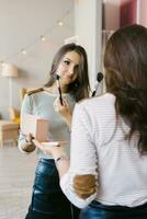 Beautiful girl puts blush on her face with a makeup brush while standing at home in a room and looking in the mirror. Caucasian girl in a striped shirt does her own makeup photo