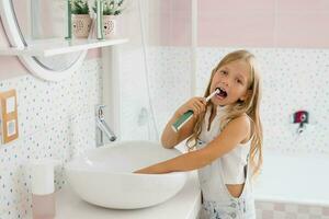 linda niño niña cepillado su dientes en frente de el baño espejo en el Mañana foto