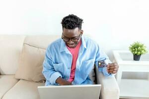 Internet payment. Happy cheerful man looking at the laptop screen while making an Internet payment. Happy young man with credit card and laptop sitting on sofa at home. Online payment photo