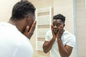 Mañana higiene, hermoso hombre en el baño mirando en espejo. reflexión de hombre africano con barba mirando a espejo y conmovedor cara en baño aseo foto