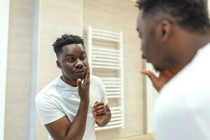 Mañana higiene, hermoso hombre en el baño mirando en espejo. reflexión de hombre africano con barba mirando a espejo y conmovedor cara en baño aseo foto