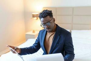 Serious Arican business man sitting on hotel bed working, analyzes expenses on business trip at the hotel room. photo