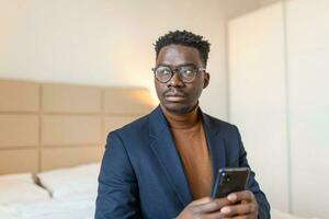 africano empresario con teléfono inteligente en un hotel habitación. hermoso hombre en pie a el ventana, enviar mensajes de texto cerca arriba. foto