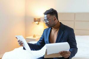 Serious Arican business man sitting on hotel bed working, analyzes expenses on business trip at the hotel room. photo