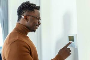 hombre afroamericano sonriente usando un moderno sistema doméstico inteligente, controlador en la pared, joven positivo cambiando la temperatura en el termostato o activando la alarma de seguridad en el apartamento foto