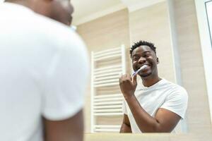 africano hombre con cepillo de dientes limpieza dientes y mirando espejo en el baño. hermoso joven hombre cepillado su dientes en Mañana en baño. contento chico cepillado dientes a noche antes de yendo a dormir. foto