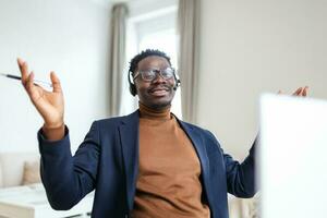 Smiling African American employee in headphones using laptop, looking at screen, making video call or watching webinar, writing notes, distance learning language concept, call center operator working photo