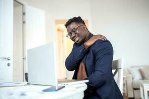 Tired African businessman working from home on his computer, feeling pain in his shoulder. man suffering from shoulder and back pain while sitting and working from home on laptop. photo