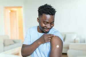 retrato de un hombre sonriente después consiguiendo un vacuna. africano hombre participación abajo su camisa manga y demostración su brazo con vendaje después recepción vacunación. foto