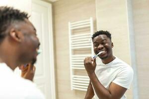 sonriente africano hombre con cepillo de dientes limpieza dientes y mirando espejo en el baño. hombre cepillado su dientes en Mañana en baño. chico en pijama cepillado dientes a noche antes de yendo a dormir. foto