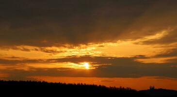 Orange sunset sky and dark purple clouds over silhouettes of monastery on the hill and forest. The sun's bright disk is partially obscured between long clouds. Diverging rays from the sun. photo