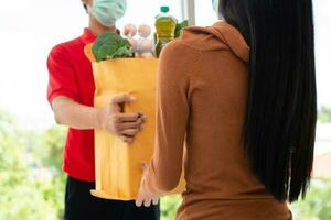 Asian delivery man from supermarket wearing a face mask and holding a bag of Fresh food, vegetables, and fruits for giving to customers at home. Concept of express grocery service and new  lifestyle photo