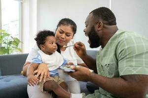 Asian mother feeding her 9 months old her cute little baby and African American helping for holding food plate At Home. Photo series of family, kids and happy people concept. Parents feed kids.