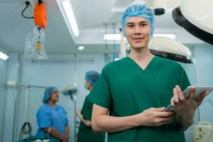 retrato de asiático cirujano con médico máscara en pie y participación un tableta en operación teatro a un hospital. equipo de profesional cirujanos cuidado de la salud, emergencia médico Servicio concepto foto