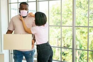 Mixed Race couple American African and Asian a surgical mask is carrying cardboard boxes in a new house on moving day. Concept of relocation, rental, and homeowner moving at home. photo