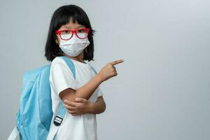 retrato de asiático niña niño con protector cara máscara con colegio mochila y punto el dedo. concepto de niño yendo espalda a colegio y nuevo normal estilo de vida foto