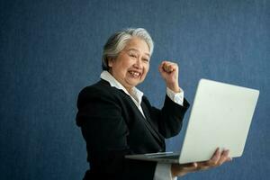 Portrait of intelligent senior businesswoman CEO manager holding laptop creating a presentation on isolated blue background. Old Woman is happy and smile after getting financial statements reports photo