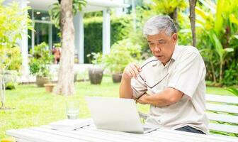 Portrait of old elderly Asian man using a computer laptop in the backyard for learning new skill after retired. Concept of no Ageism and not be late for learning. photo