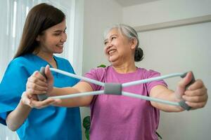 Happy elderly female exercising with rubber expander with Young physiotherapist for recovery arm muscles in a physiotherapy center. Concept of happy retirement with care from a caregiver and health. photo