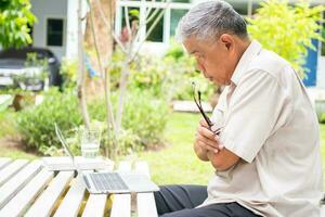 Portrait of old elderly Asian man using a computer laptop in the backyard for learning new skill after retired. Concept of no Ageism and not be late for learning. photo