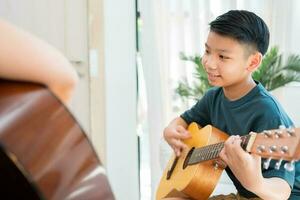 Asian boy playing guitar with father in the living room for teaching him son play guitar, feel appreciated and encouraged. Concept of a happy family, learning and fun lifestyle, love family ties photo