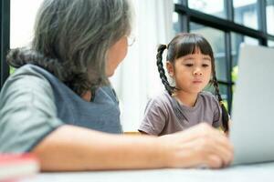 abuela asiática con sus dos nietos divirtiéndose y jugando juegos educativos en línea con un ordenador portátil en casa en la sala de estar. concepto de educación en línea y cuidado de los padres. foto