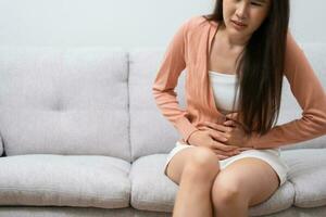 Sick Asian Woman Suffering From Acute Abdominal Pain in the abdomen due to menstruation period, PMS.  Sitting On Couch, stomachache from food poisoning, abdominal pain, digestive problem, gastritis photo