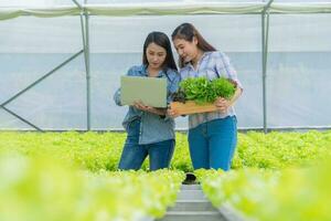 mujer agricultora asiática que sostiene una cesta de verduras de ensalada de verduras frescas en una granja orgánica y usa una computadora portátil para verificar el pedido del cliente. concepto de agricultura orgánica para la salud y pequeñas empresas. foto