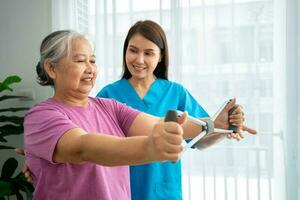 Happy elderly female exercising with rubber expander with Young physiotherapist for recovery arm muscles in a physiotherapy center. Concept of happy retirement with care from a caregiver and health. photo