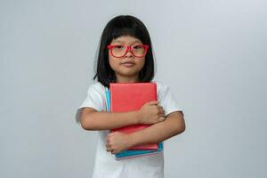 Happy Asian little preschool girl wearing red glasses holding books and red backpack on white isolated background. Concept of school kid and education in elementary and preschool, home school photo