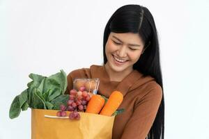 contento asiático mujer es sonriente y lleva un compras papel bolso después el mensajero desde el tienda de comestibles llegó a entregar su bienes a hogar. concepto de supermercado entrega para un nuevo estilo de vida foto