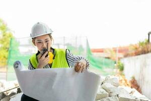 Asian engineer or Young woman Architect put on a helmet for safety and look at Blueprint for Inspect Building factory Construction Site and use walkie talkie for a talk with a contractor. photo