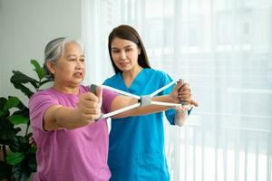 Happy elderly female exercising with rubber expander with Young physiotherapist for recovery arm muscles in a physiotherapy center. Concept of happy retirement with care from a caregiver and health. photo