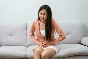 Sick Asian Woman Suffering From Acute Abdominal Pain in the abdomen due to menstruation period, PMS.  Sitting On Couch, stomachache from food poisoning, abdominal pain, digestive problem, gastritis photo