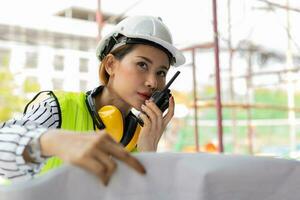Asian engineer or Young woman Architect put on a helmet for safety and look at Blueprint for Inspect Building factory Construction Site and use walkie talkie for a talk with a contractor. photo