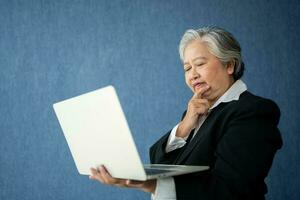 Portrait of intelligent senior businesswoman CEO manager holding in hands laptop creating a presentation on isolated blue background. Old Woman thinking after getting financial statements reports. photo