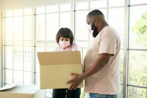 Mixed Race couple American African and Asian a surgical mask is carrying cardboard boxes in a new house on moving day. Concept of relocation, rental, and homeowner moving at home. photo