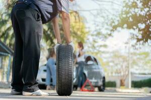 Expertise mechanic man in uniform holding a tire for help a woman for changing car wheel on the highway, car service, repair, maintenance concept. photo