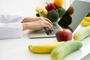 Portrait of Asian smiling female nutritionist typing on laptop computer for balanced corrective diet plan for patient or weight loss advice. Concept of healthy lifestyle, healthy food, dieting photo